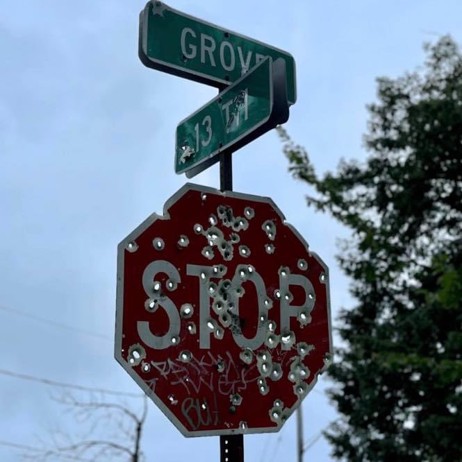 A weathered stop sign riddled with bullet holes, located at the intersection of Grove Street and 13th Avenue.