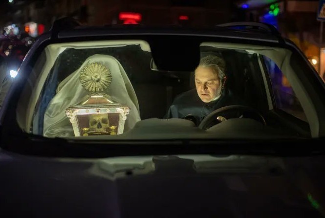 A man sitting in a car at night, focused on his task while a religious relic, covered with a veil and adorned with a sunburst design, is visible in the passenger seat.