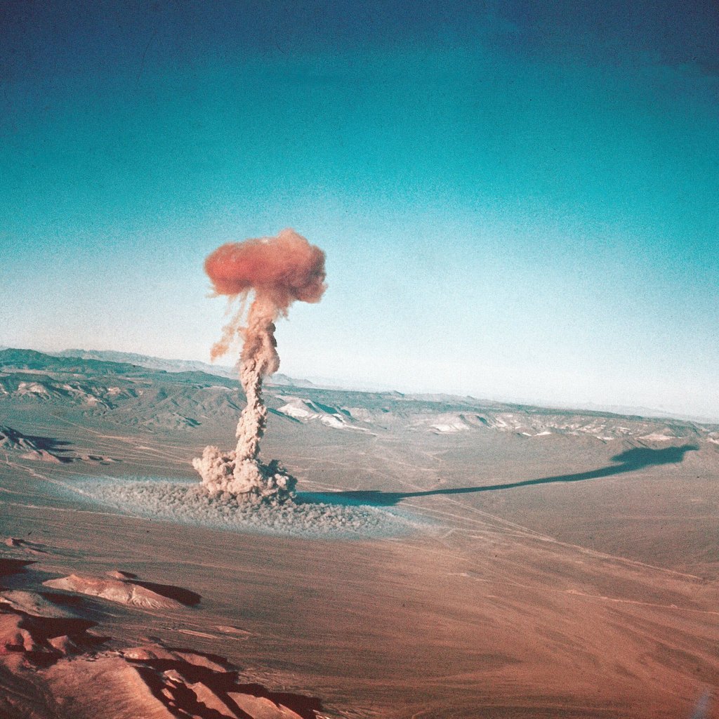 Aerial view of a large mushroom cloud rising above a desert landscape, with a blue sky in the background.